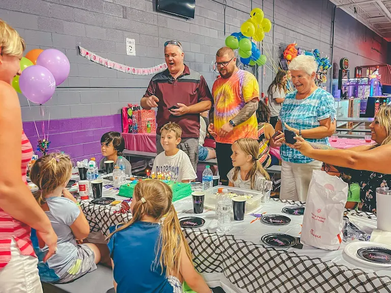 Group celebrating a joyful birthday party with kids and adults gathered around a decorated table, featuring balloons, gifts, and a festive atmosphere.